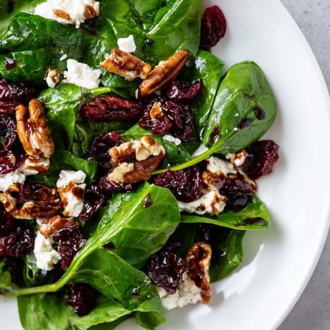 The spinach goat cheese salad features emerald spinach leaves, ruby cranberries, and crunchy candied pecans in a glossy balsamic dressing on a white plate.