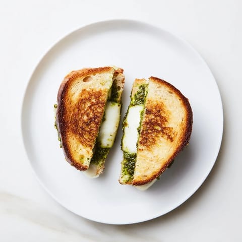 A close-up of a sourdough pesto grilled cheese, revealing layers of sharp white cheddar, fontina, and aromatic basil pesto between toasted slices.